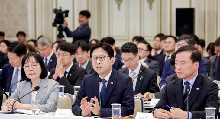 Government ministers seated at a policy briefing table, one speaking while others listen