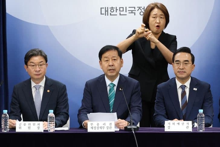Government economic officials seated at a podium during a joint policy briefing, with a sign language interpreter behind them