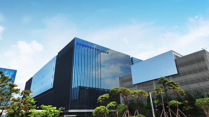 The Samsung Biologics headquarters building, a dark glass facade photographed from below against a blue sky